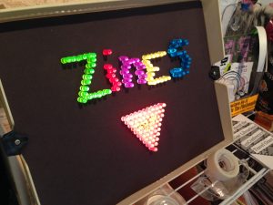 Lite-Brite board displaying the word "Zines" and a pink triangle, on display at QZAP.