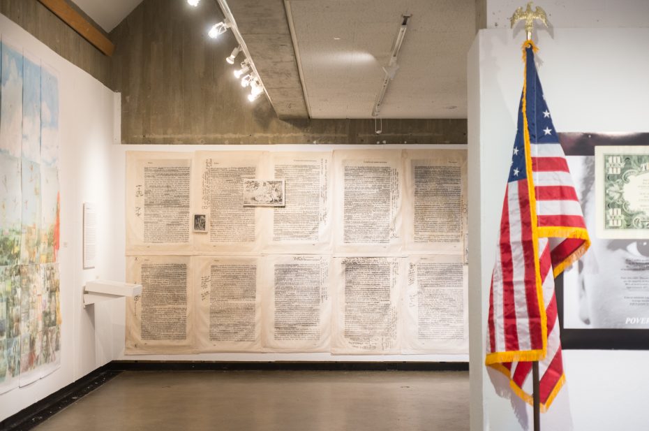 photograph of two gallery walls, one with blown up fabric prints of annotated book pages, and an american flag against another gallery wall