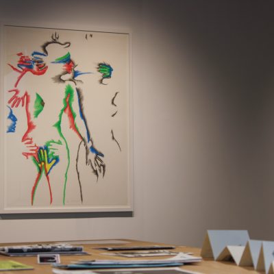 One of six lithographs in a series by the artist Marisol hangs on a gallery wall depicting intimately entangled human subjects in bright color while a wooden table holding zines stands in the foreground
