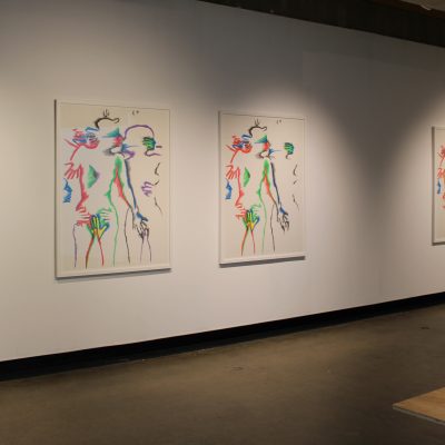 Behind the edge of a wooden table holding zines a set of six lithographs by the artist Marisol depicting intimately entangled human subjects in increasing color hang on a wall in groupings of three shown at a slight angle from the viewer