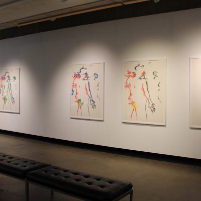 Behind two benches a set of six lithographs by the artist Marisol depicting intimately entangled human subjects in increasing color hang on a wall in groupings of three shown at a slight angle from the viewer