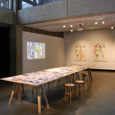 A wooden table holding zines stands parallel to a projection of the tabletop and perpendicular to three lithographs by the artist Marisol which hang on a gallery wall in the background