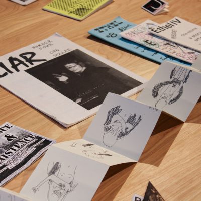 A collection of zines laid out on a wooden tabletop with one zine being an accordion style series of drawings and with visible titles including LIAR and PERSISTENCE & EXISTENCE and Ethel IV and ETHEL #8