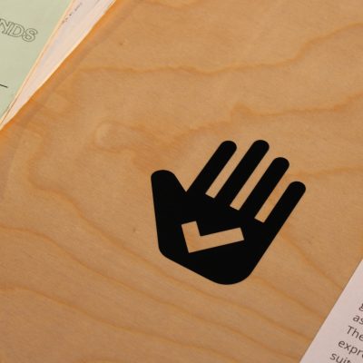 A black vinyl sticker of a hand with a checkmark cut out of its palm is shown on a wooden tabletop with zines and descriptive text partially visible on either side