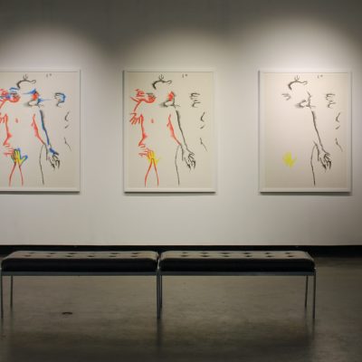 A set of three lithographs by the artist Marisol depicting intimately entangled human subjects in increasing color hang on a wall behind two benches and face the viewer