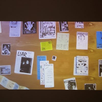 A projection shows an aerial view of zines arranged on a wooden tabletop with visible titles including LIAR and Mohawk Beaver