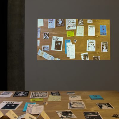 A wooden table holding zines with visible titles such as LIAR stands in the foreground while a projection of zines arranged on the tabletop is shown on the wall behind