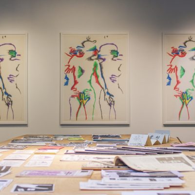 A set of three lithographs by the artist Marisol depicting intimately entangled human subjects in increasing color hang on a gallery wall facing the viewer while a wooden table holding zines stands in the foreground