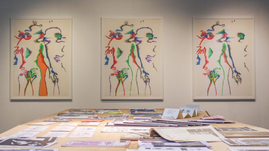 A set of three lithographs by the artist Marisol depicting intimately entangled human subjects in increasing color hang on a gallery wall facing the viewer while a wooden table holding zines stands in the foreground