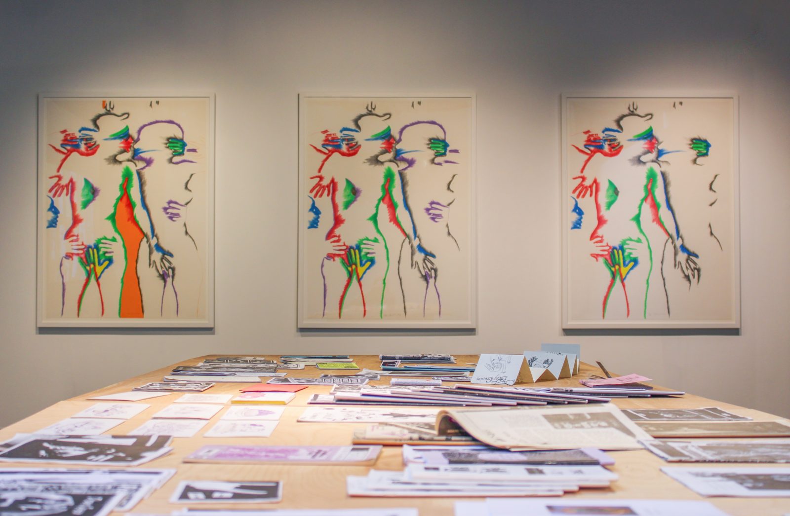 A set of three lithographs by the artist Marisol depicting intimately entangled human subjects in increasing color hang on a gallery wall facing the viewer while a wooden table holding student zines stands in the foreground