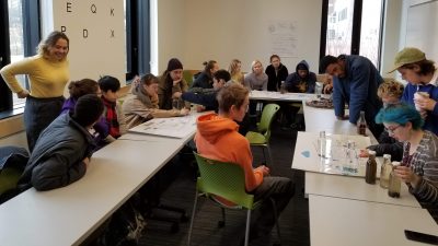 A classroom filled with college students who are all wearing winter attire. They are looking at the kern water filtering system drawn out on posters. Bottles of different filtered stages of water on the desk.