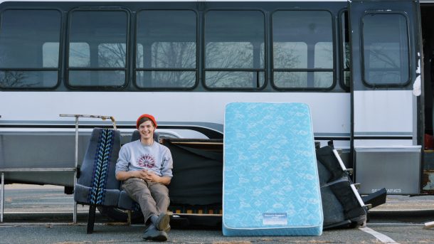 A student wearing a Hampshire College sweater sitting on bus seats that have been ripped out and places outside leaning against the bus. Next to them is a blue clean mattress that leans against more gutted bus parts.