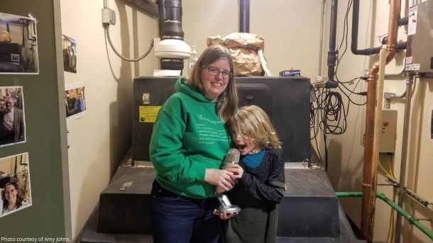 A women standing with her child. The women is wearing a green hoodie with unreadable words and blue jeans. She is holding a microphone for her child. They are in what appears to be a water filter system room.