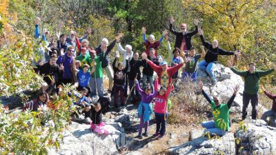 A group photo with over 15 people standing at the top of a hiking trail with both of their arms raised high. They are dressed for the autumn season.