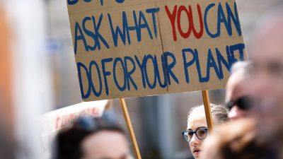 A picture with blurry people in the frame and only focusing on a person in the background with blue glasses. They are holding a cardboard poster sign that reads, "Ask what you can do for your planet," in blue paint and the word "YOU" in red paint.