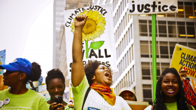 A climate change protest with black femmes in frame holding different posters and wearing green shirts with a slogan that can't be read.