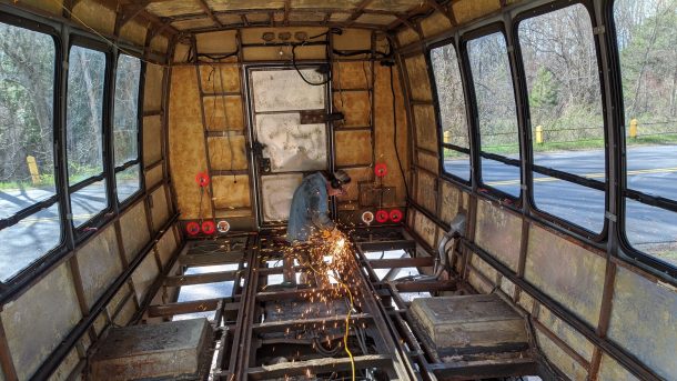 The rusty inside of a gutted bus with no floor that has a welder melting floor metal together. Sparks are flying