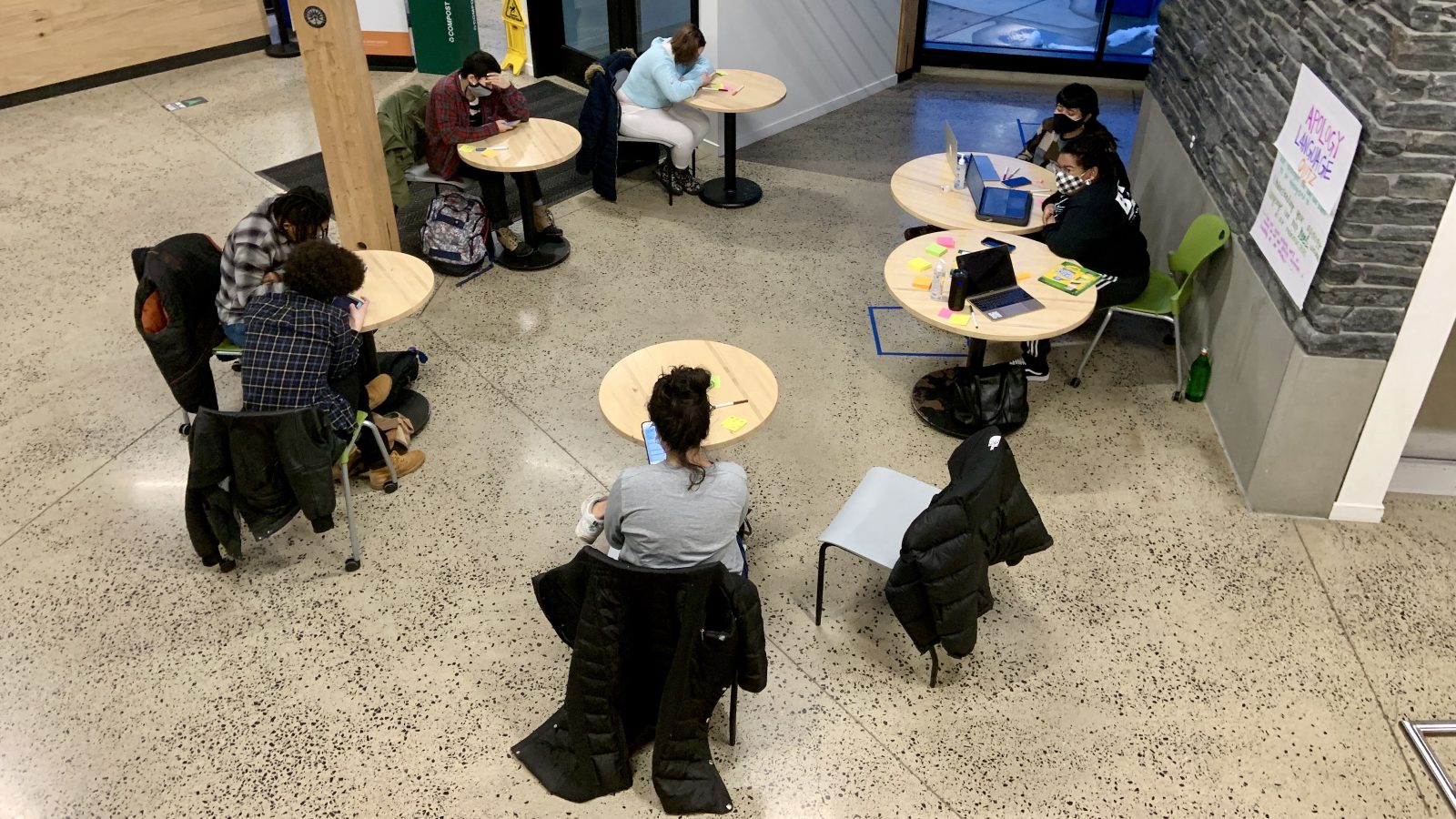 A high view looking down on students sitting at different tables looking at their phones and laptops