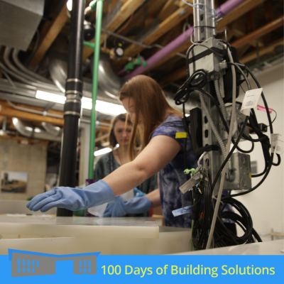 Two people stand blurred in the background over an open greywater tank. In focus in the foreground is a tangle of electrical conduits and wires that wrap their way up a metal pole. A banner at the bottom of the image includes a simple shape of the R.W. Kern Center and reads: 100 Days of Building Solutions.