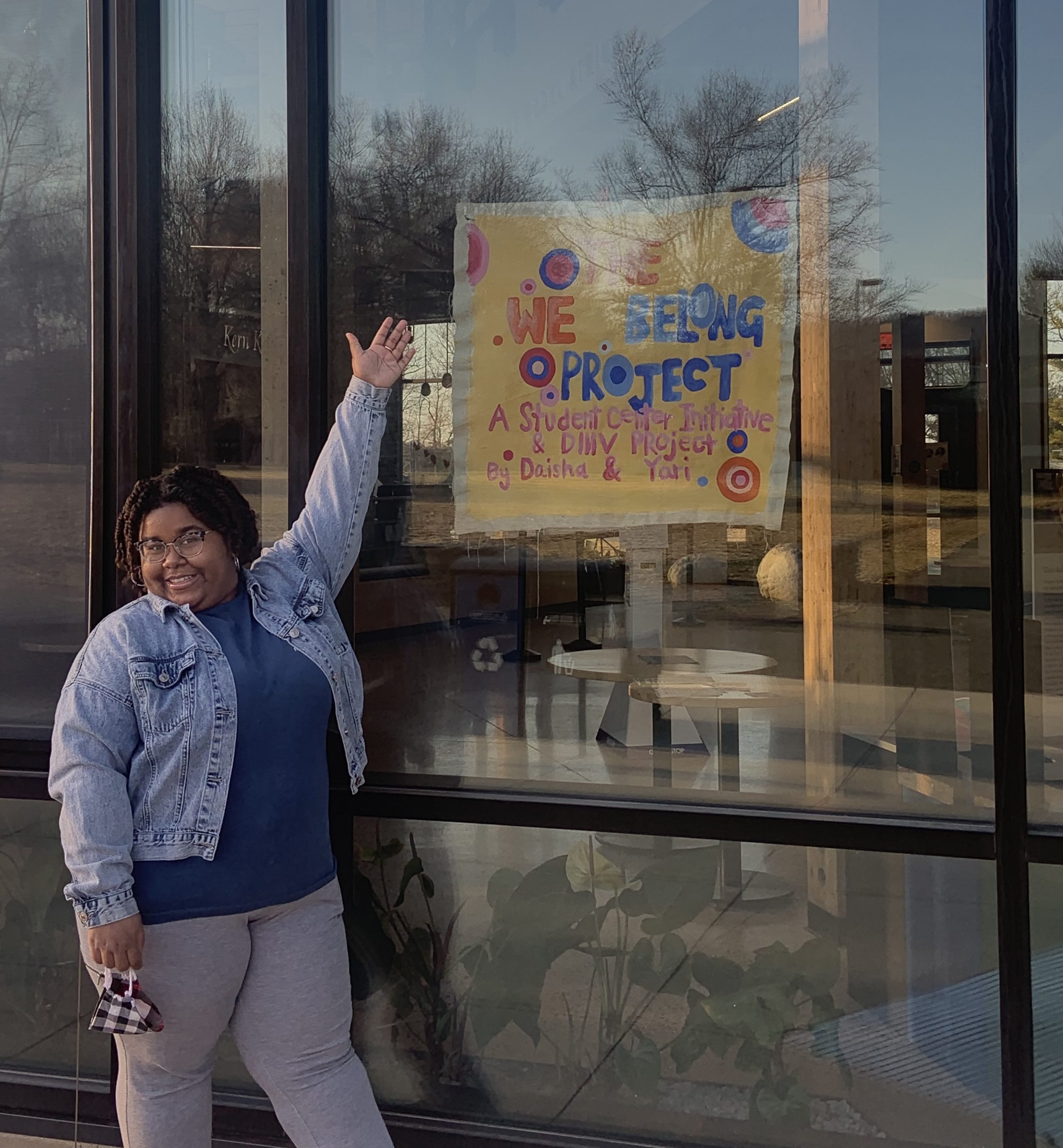 Daisha stands in front of the R.W. Kern Center gesturing toward a We Belong Project banner.