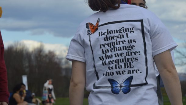 A picture focusing on a person's shirt. The shirt has a black line rectangle with a monarch butterfly in the upper left corner and a blue butterfly in the middle at the bottom. In the rectangle, it reads, "Belonging doesn't require us to change who we are; it requires us to BE who we are."