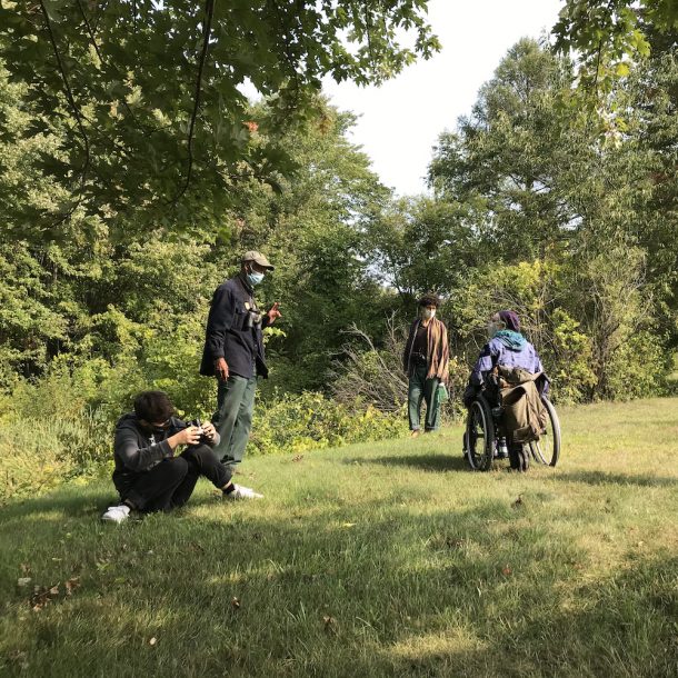 A picture of four people outside on the grass near the edge of the tree line. One person is in a wheelchair, one is sitting on the grass, and two are standing. They are all wearing autumn attire.