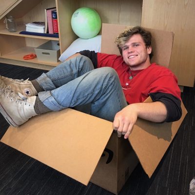 Oliver, wearing jeans and a red t-shirt, sitting in a cardboard box.