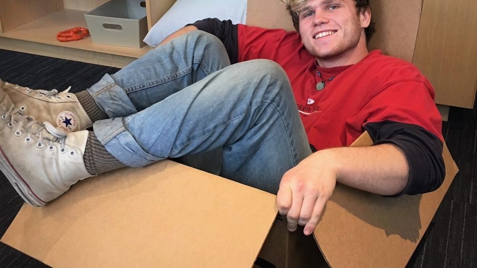 Oliver, wearing jeans and a red t-shirt, sitting in a cardboard box.